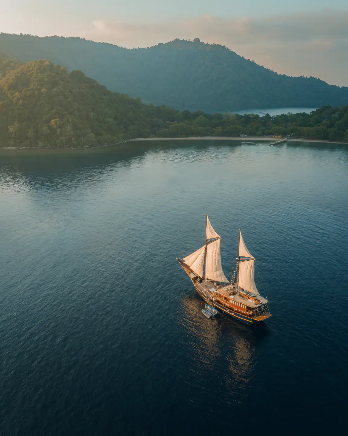 Aerial of Celestia at anchor with sails set in a calm forested bay