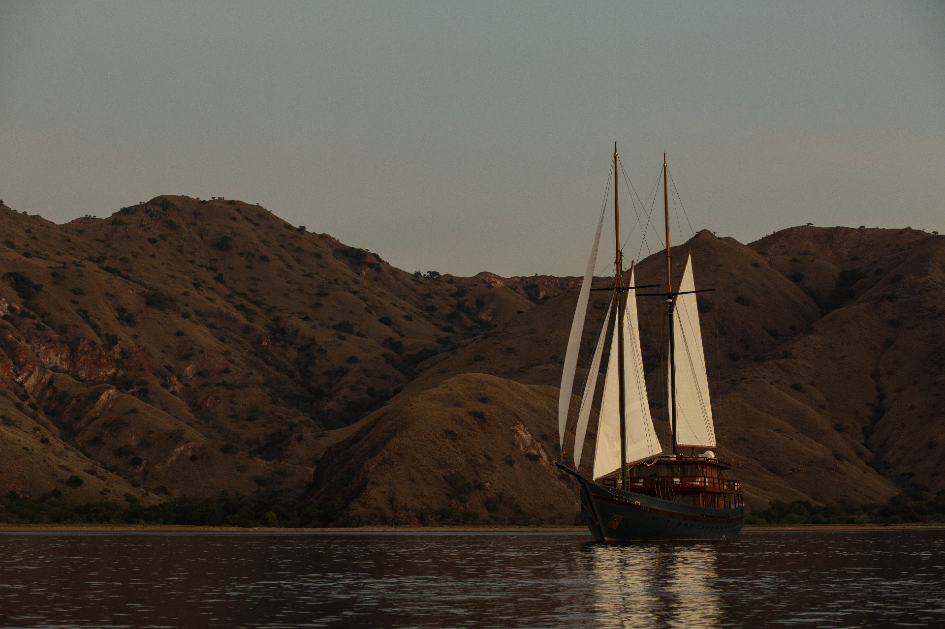 Sailing yacht Vela anchored under full sail at sunset in front of Komodo hills