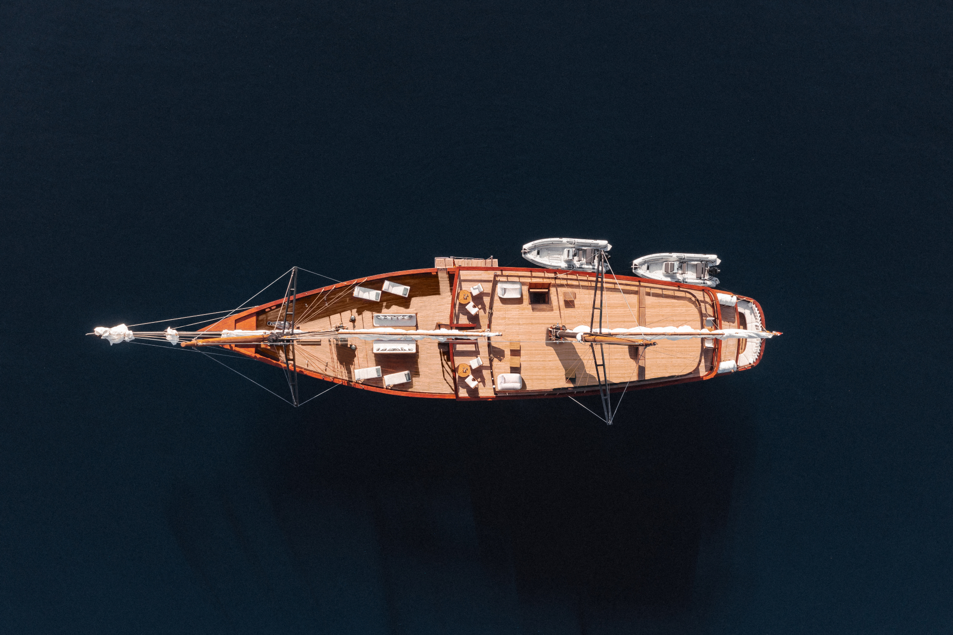 Aerial top-down of Vela on deep navy water showing teak decks, sunloungers and two tenders stowed aft