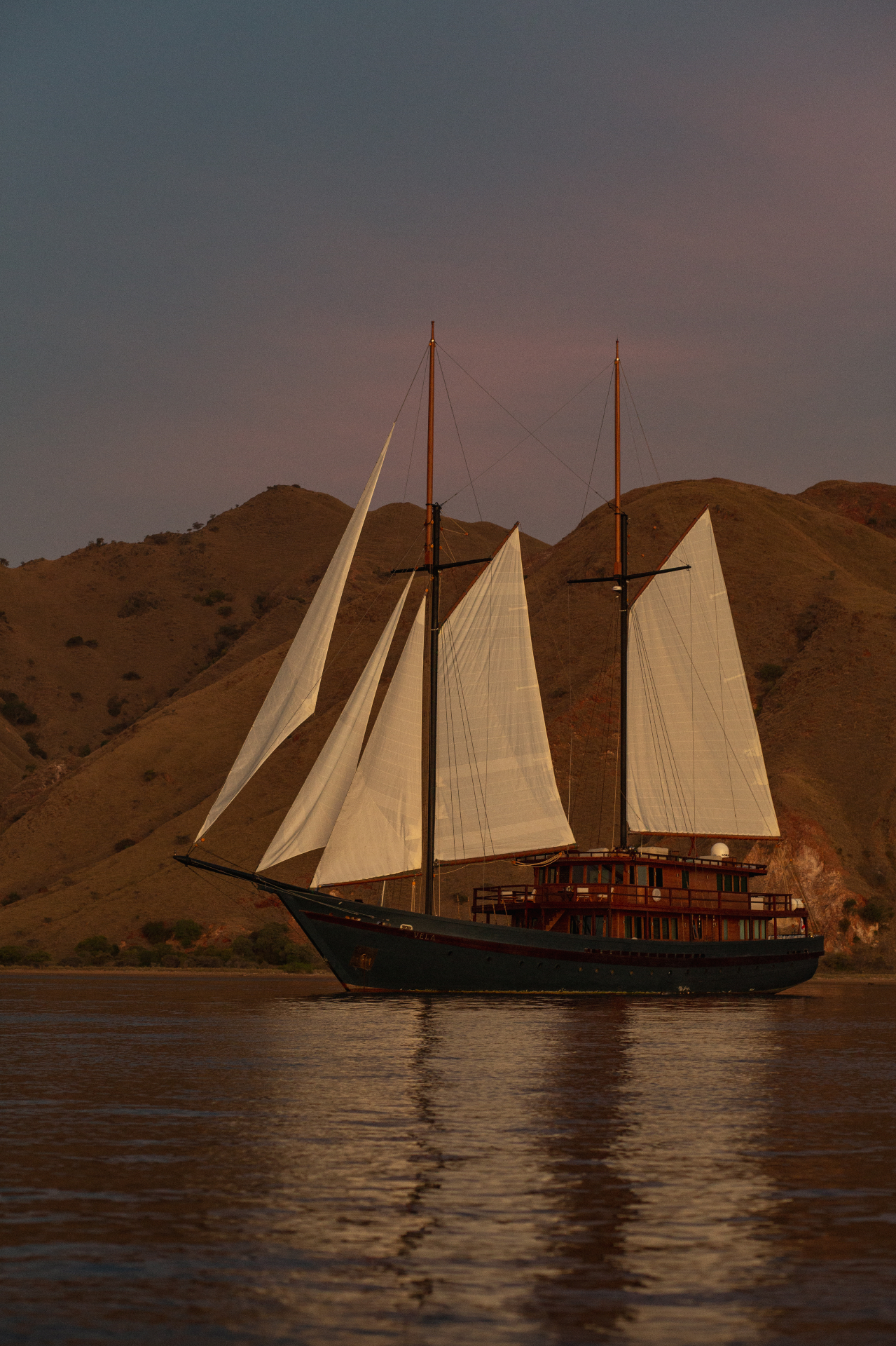 Vela's twin masts and white sails fully rigged at dusk with pink sky