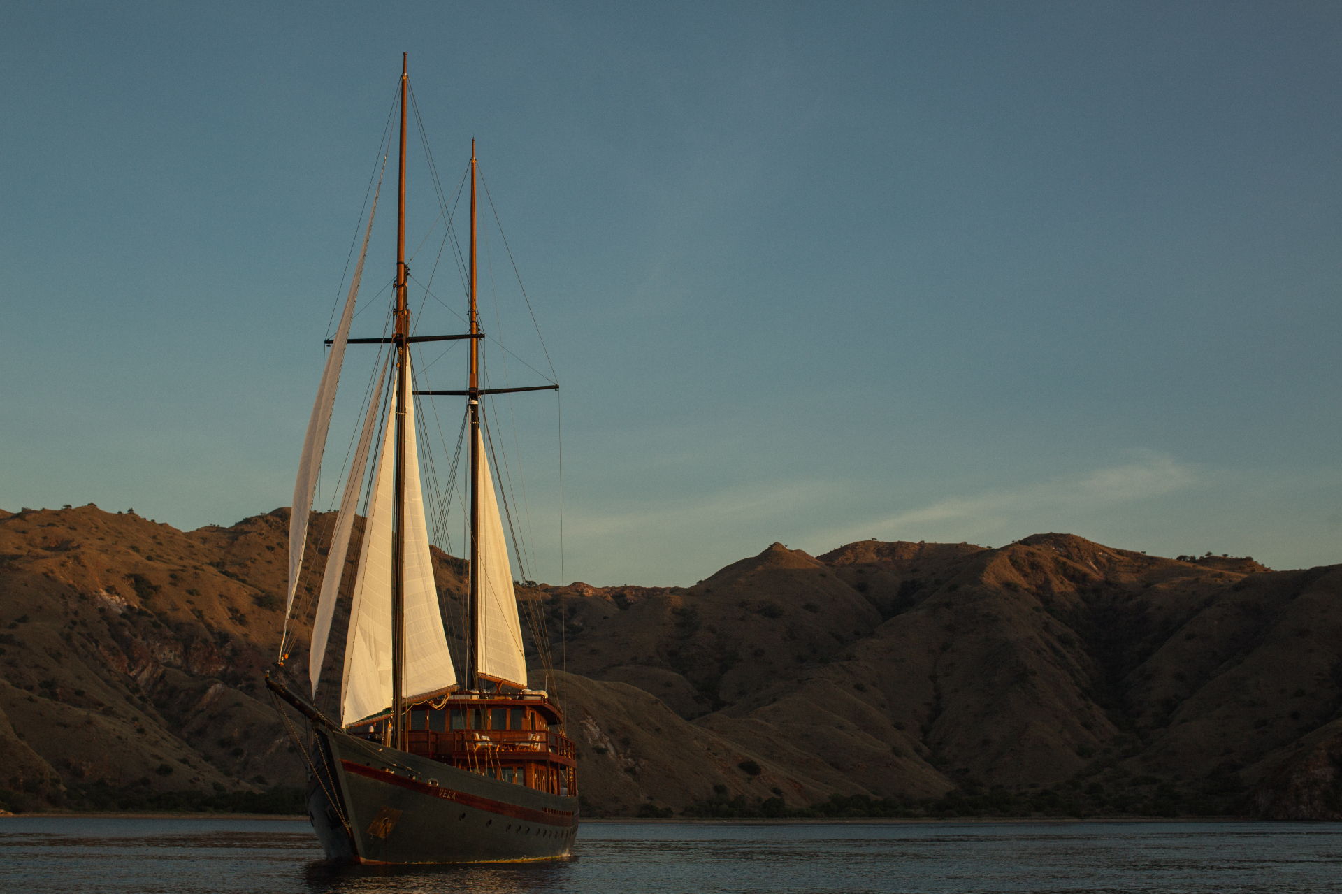 Vela sailing on calm water with twin masts and white sails, mountainous island behind
